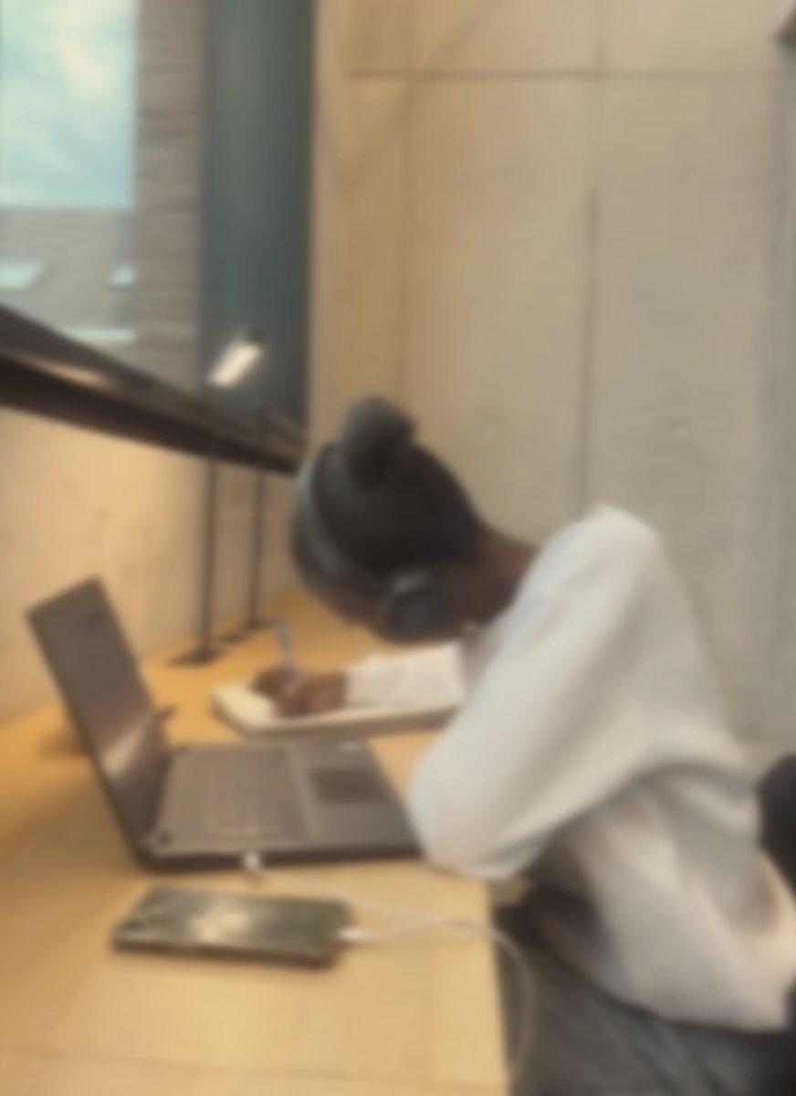 A student in headphones writing in a notebook beside a laptop